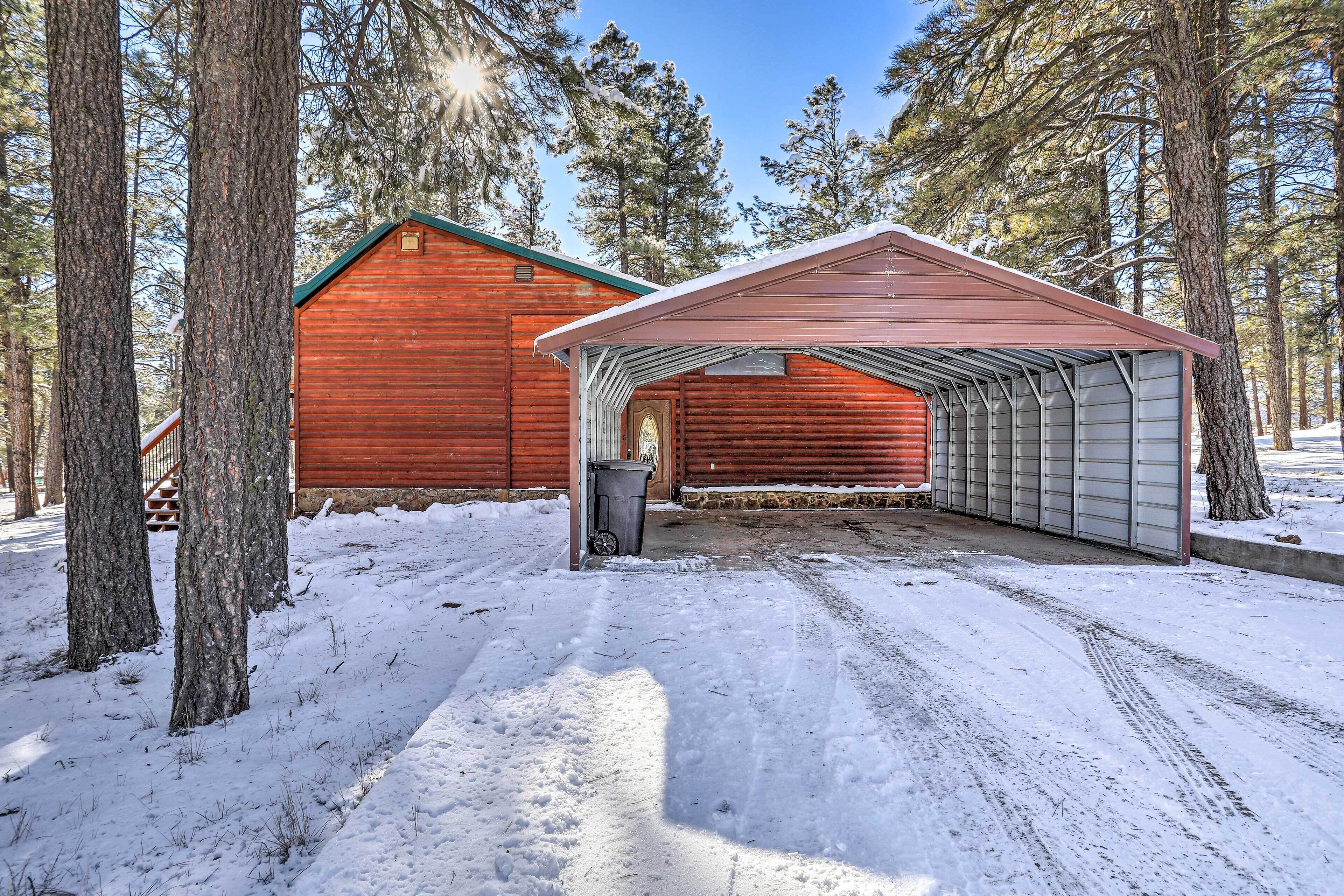 A wooden cabin with a metal garage and a covered porch, surrounded by tall trees and snow.