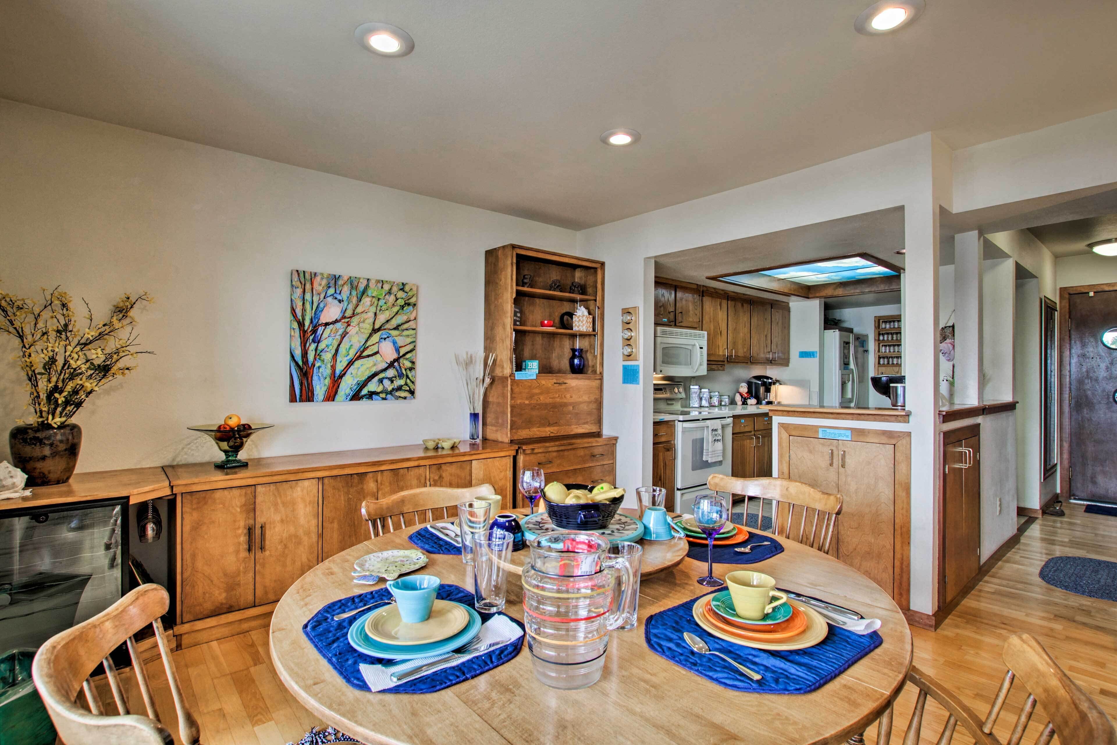 A dining area with a round table set for four, wooden cabinets, a kitchen in the background, and a painting on the wall.