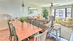 A spacious living area with a wooden dining table, comfortable seating, a large mirror, and a view of the city through large windows. - Summer Winds Resort Condo w/ Ocean Views! (Atlantic Beach)