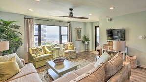 A spacious living room with a large sofa, a coffee table, and a television. There is a large window with a view of the ocean. - Summer Winds Resort Condo w/ Ocean Views! (Atlantic Beach)