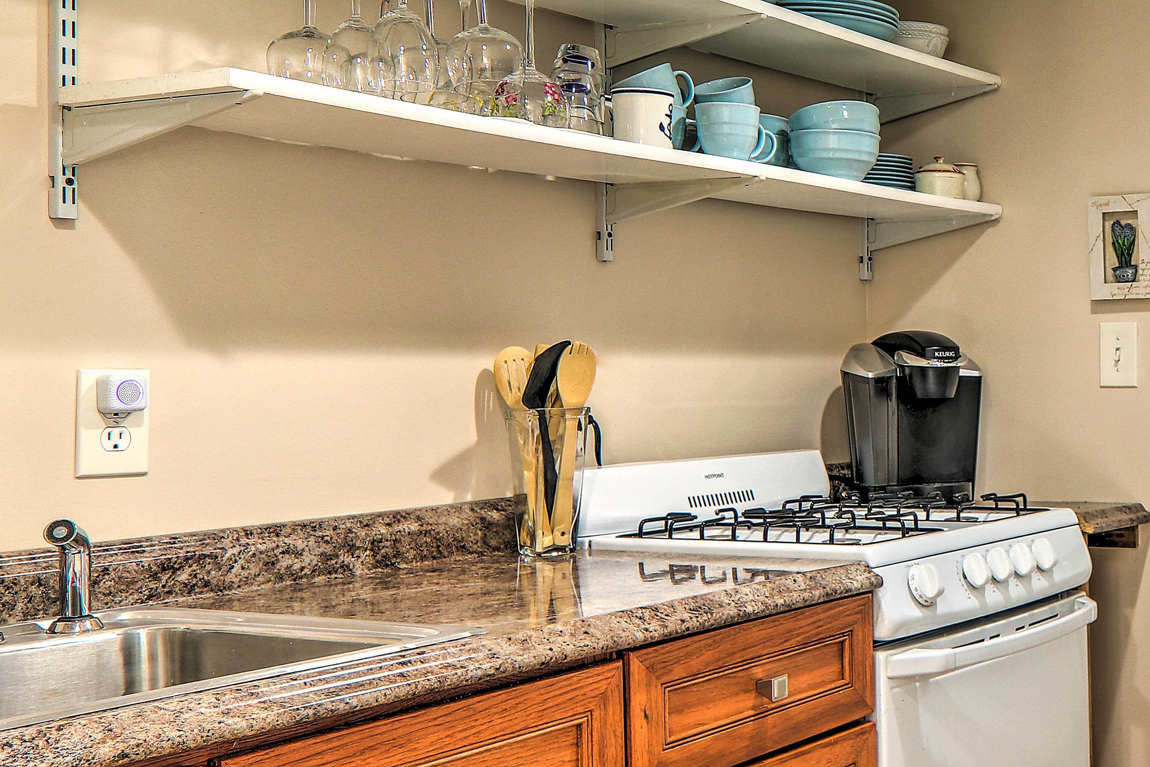 A kitchen with a gas stove, a coffee maker, a sink, and shelves above holding dishes and glasses.