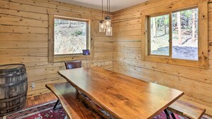 A wooden cabin interior with a large dining table, two windows, a hanging light fixture, and a wooden barrel.