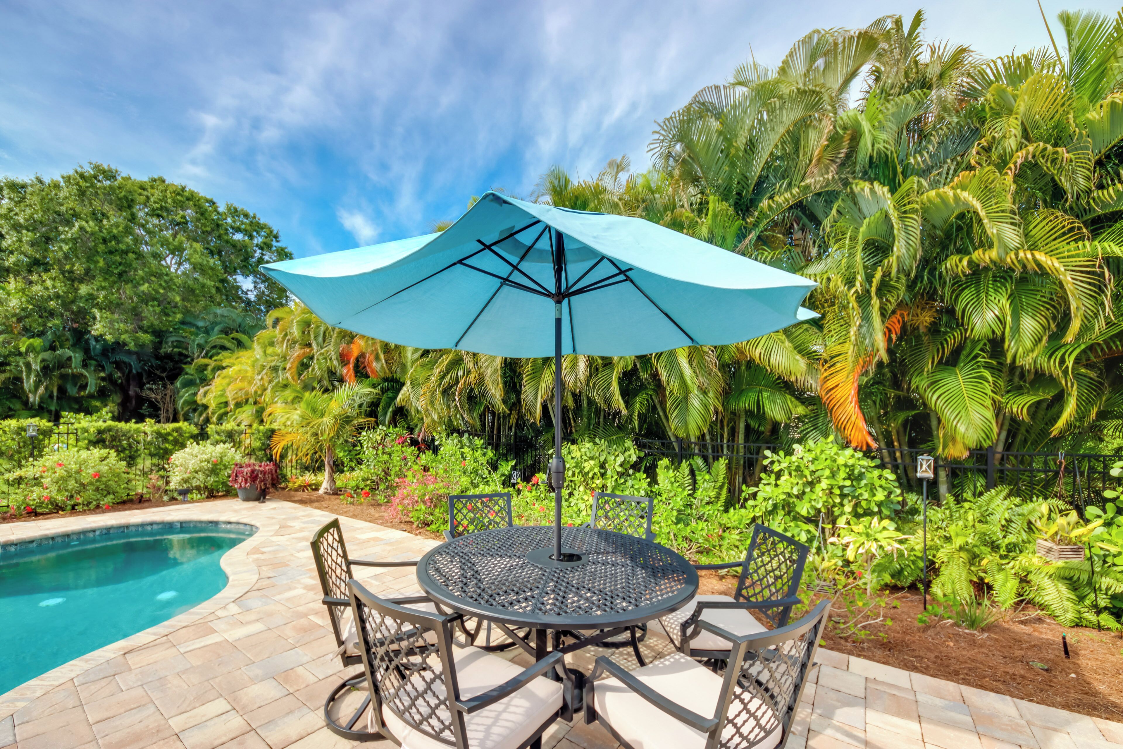 Une table ronde placée à l’extérieur, surmontée d’un parasol bleu, entourée de chaises, près d’une piscine, avec une végétation luxuriante et des palmiers en arrière-plan.