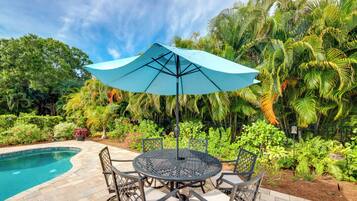 Une table ronde placée à l’extérieur, surmontée d’un parasol bleu, entourée de chaises, près d’une piscine, avec une végétation luxuriante et des palmiers en arrière-plan.