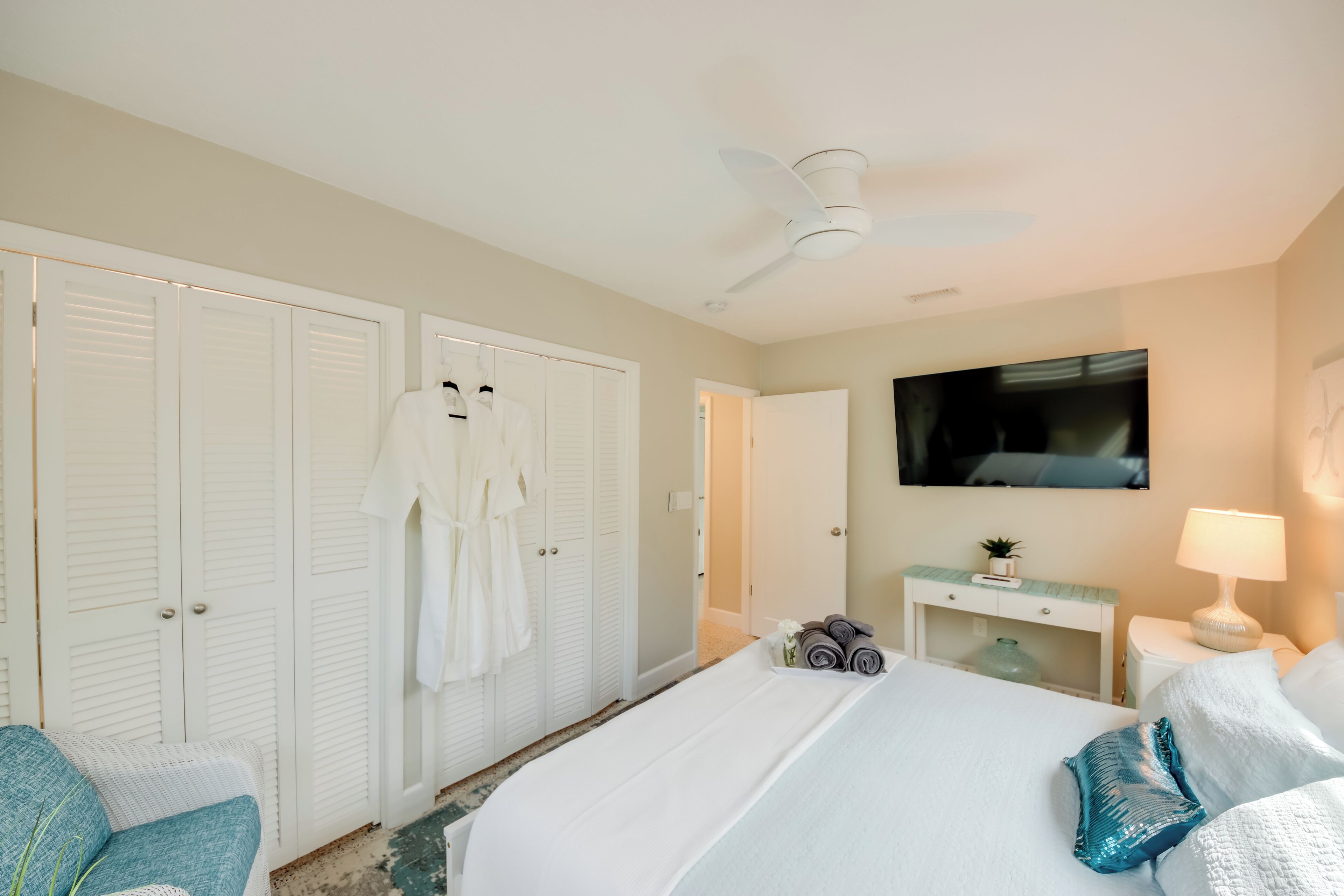 A hotel room with a bed, a TV, a ceiling fan, a desk with a lamp, and a wardrobe.