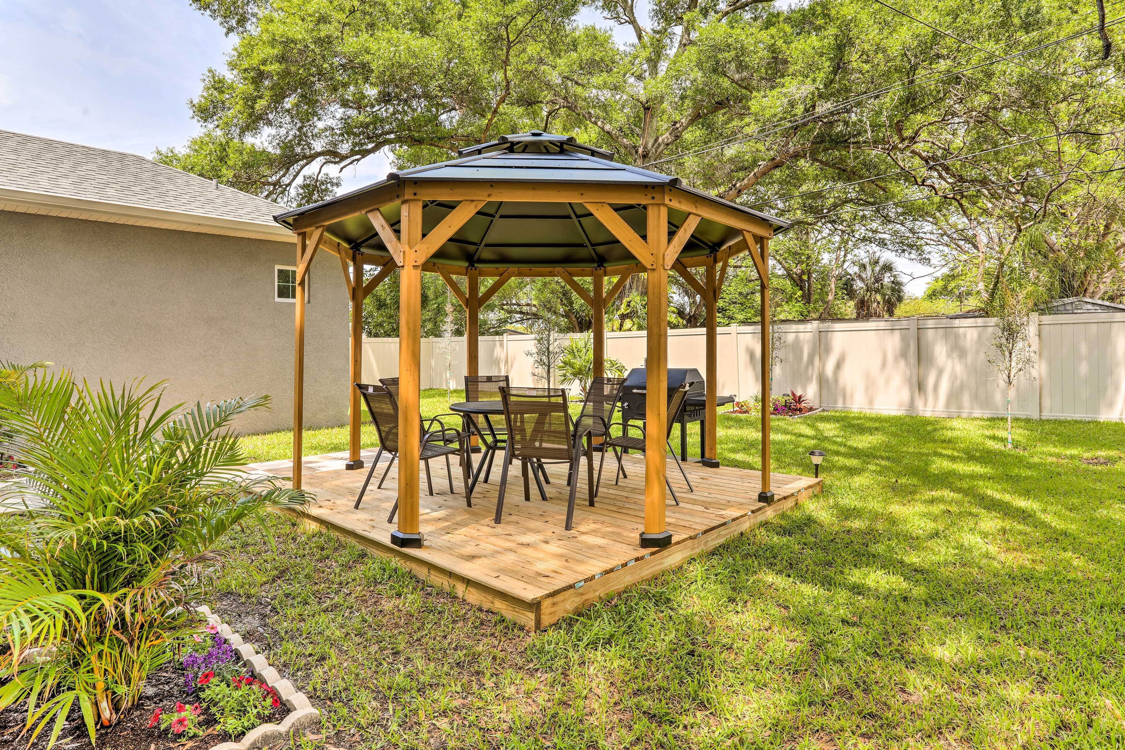 A wooden gazebo with a roof, surrounded by a grassy yard and a wooden deck.
