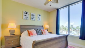 Quarto com cama de madeira, duas mesas de cabeceira com luminárias, uma janela ampla com vista para palmeiras e três quadros na parede.