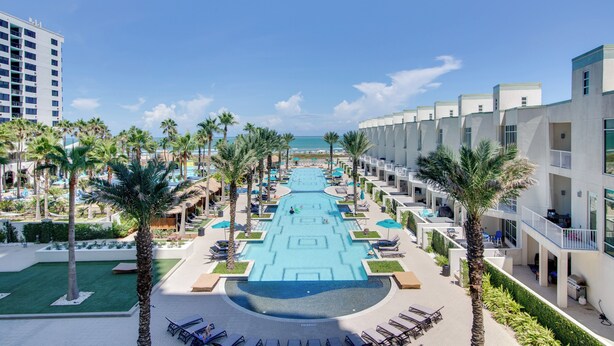 Um resort com piscina central, palmeiras e vista para o oceano.
