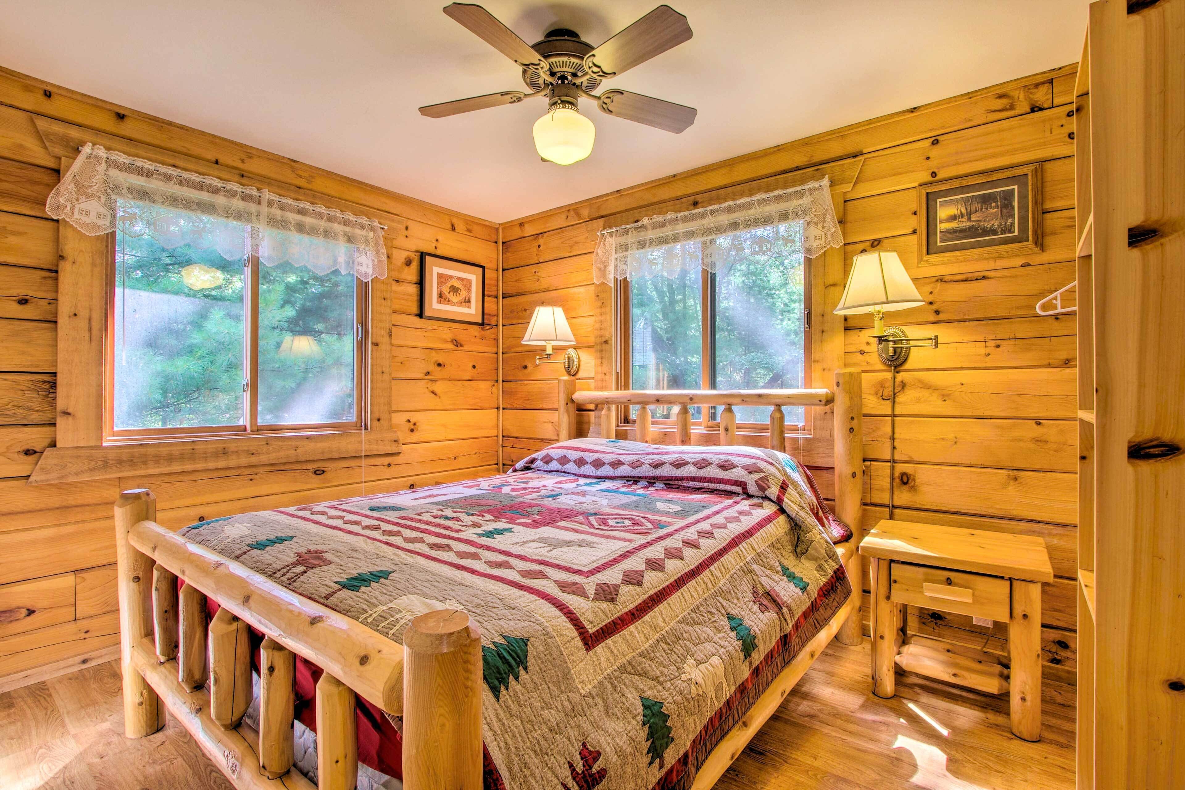 A wooden cabin bedroom with a bed, two windows with lace curtains, a ceiling fan, and a small table with a lamp.