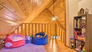 A cozy attic room with wooden beams, a sloped ceiling, a ceiling fan, bean bags, a bookshelf, and a window. - Pet-friendly Warrens Cabin w/ Fire Pit! (Warrens)
