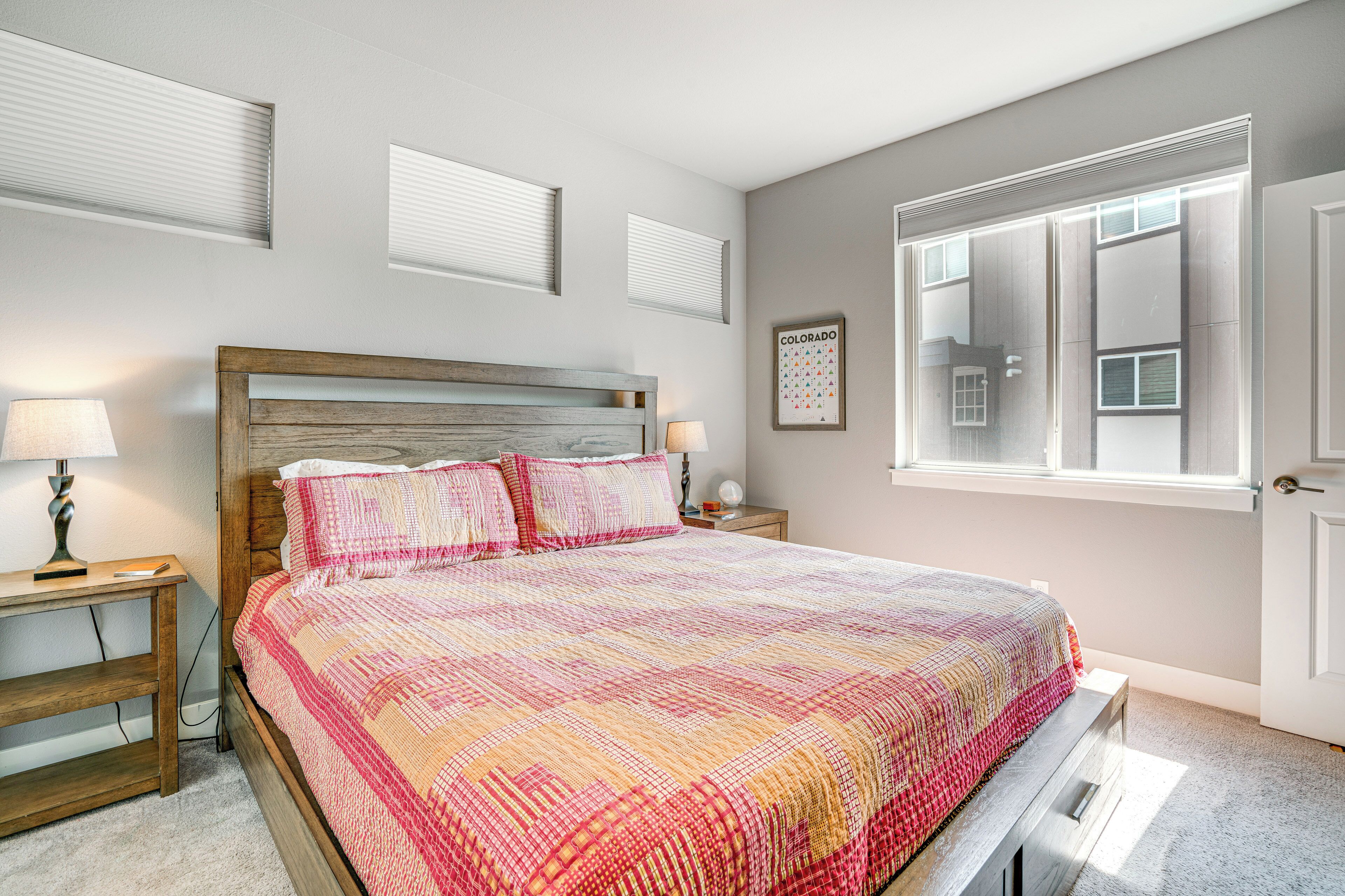 Une chambre à coucher avec une tête de lit en bois, un grand lit recouvert d’une couette aux couleurs vives, des lampes de chevet, une table de chevet et une fenêtre avec des stores.