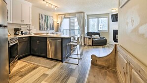Eine moderne Küche mit Kochinsel, weißen Schränken und Edelstahlgeräten. Ein Essbereich mit einer Couch und einem an der Wand befestigten Fernseher.