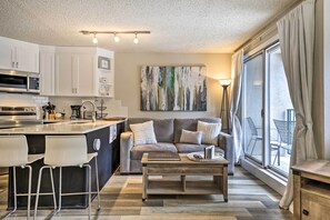A modern kitchen with white cabinets, a built-in microwave, and a bar area with white stools. There is a grey sofa with cushions, a wooden coffee table, and a large window with curtains. - Ski-in/ski-out Winter Park Condo w/ Balcony! (Winter Park)