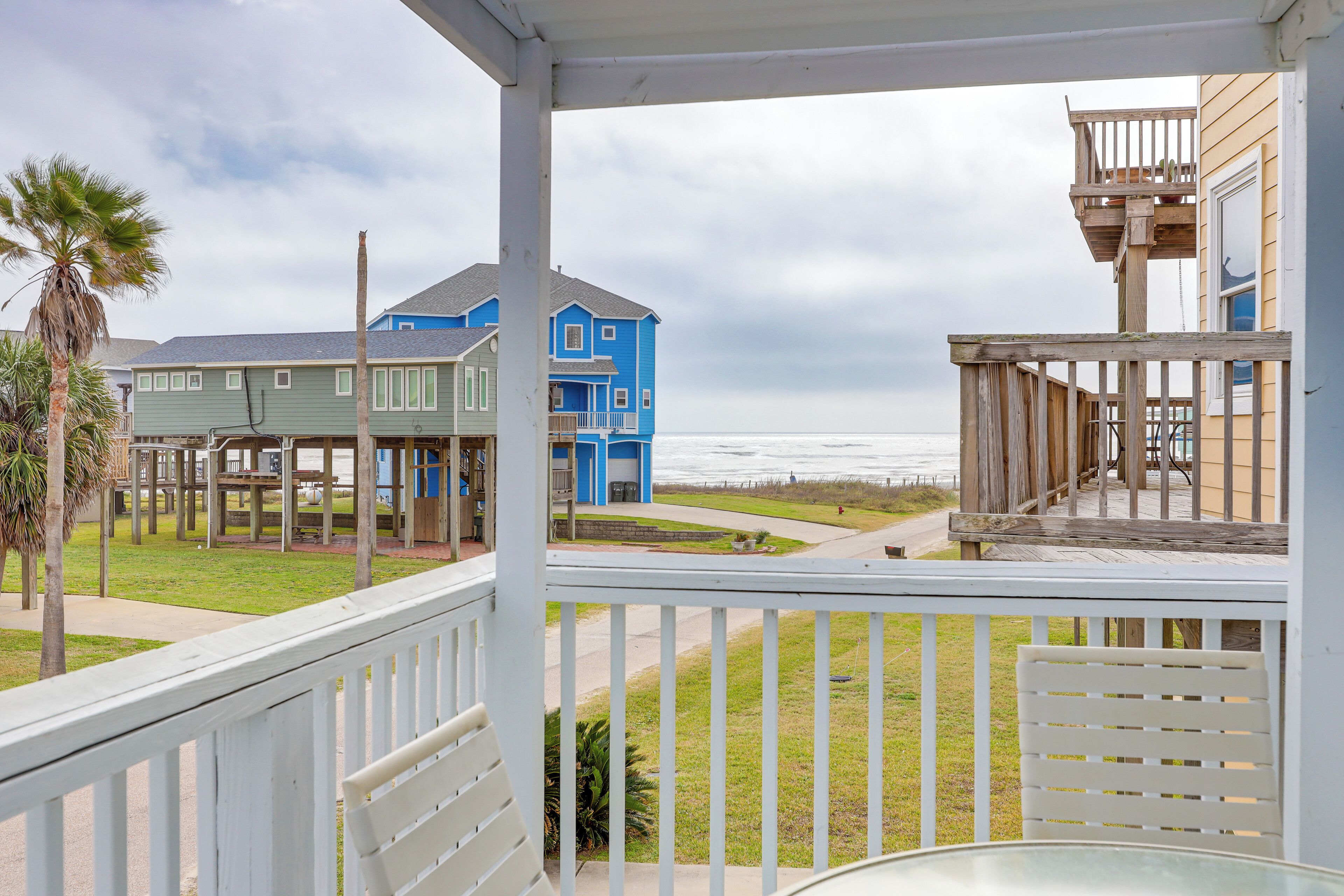 A coastal view from a balcony with a wooden railing, overlooking a beach and a blue house with a wooden deck.