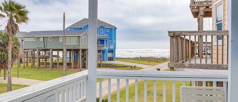 A coastal view from a balcony with a wooden railing, overlooking a beach and a blue house with a wooden deck.
