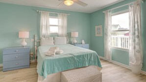 A bedroom with teal walls, a bed with white bedding, two bedside tables with lamps, a ceiling fan, and a window with curtains. - Steps to Beach: Galveston Vacation Rental! (Galveston)