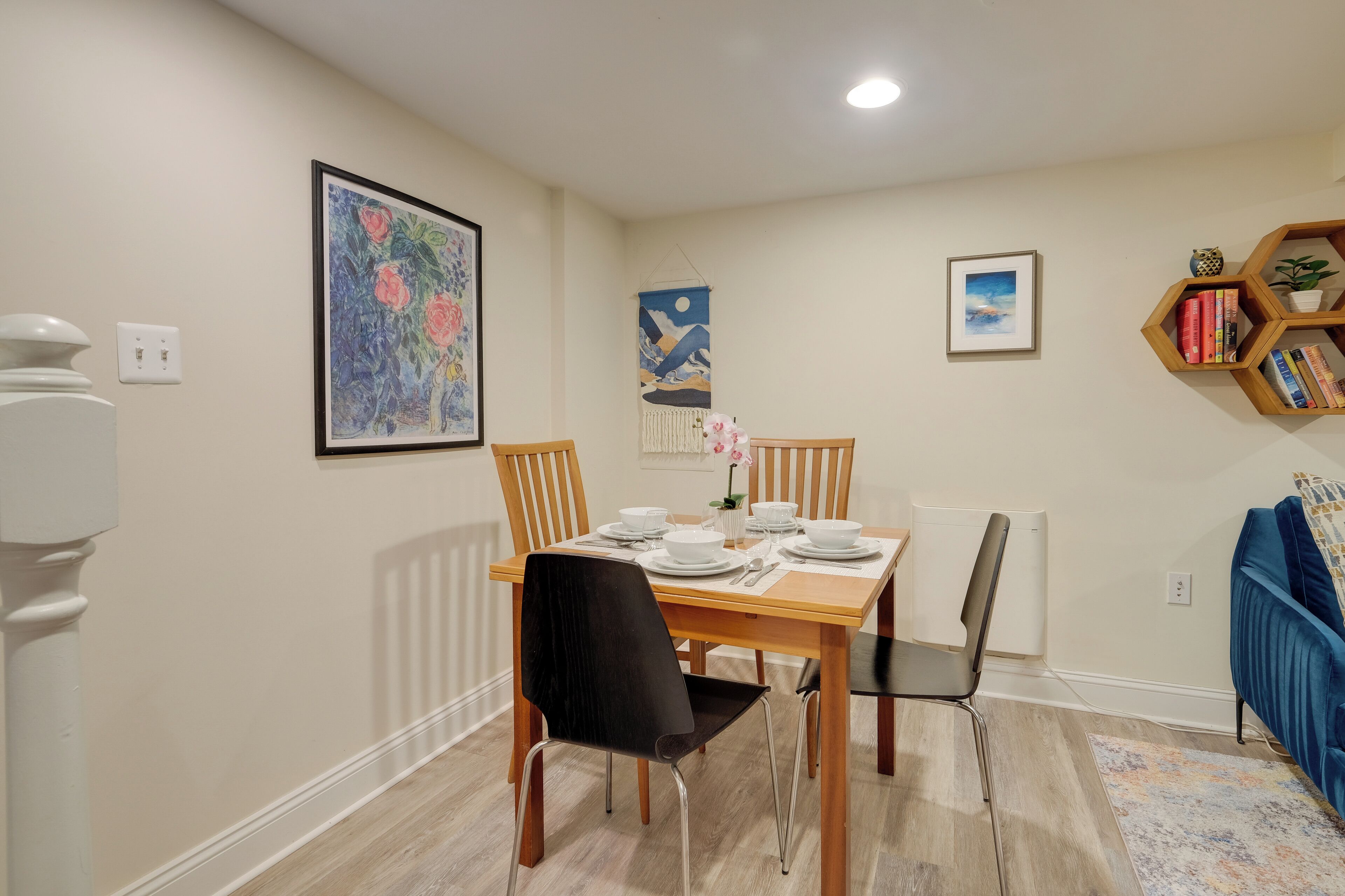 A dining area with a wooden table set for four, surrounded by chairs. There are framed artworks on the walls, a radiator, and a bookshelf with books and decorative items.