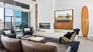 A modern living room with a large TV, comfortable seating, a surfboard, and a view of the ocean.