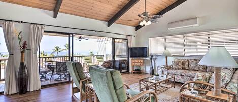A spacious living room with wooden flooring, a ceiling fan, a flat-screen TV, wicker furniture, and large windows offering a view of palm trees.