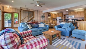 A spacious living room with blue sofas, a wooden coffee table, and a checkered ottoman. A staircase leads to a balcony with a view of trees. - Sunny 'providence Cottage' Retreat w/ Dock, Kayaks (Sunrise Beach)