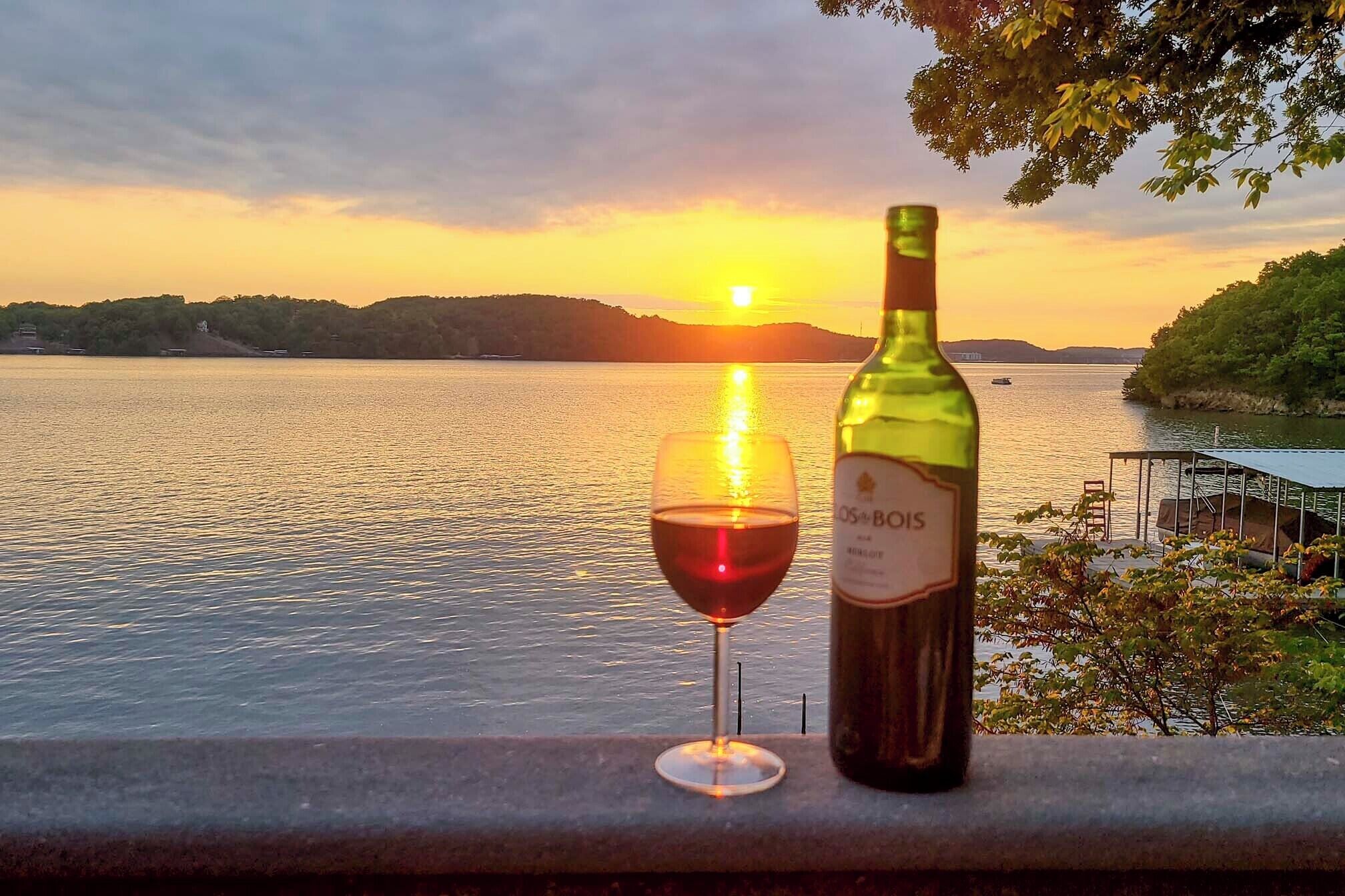 A bottle of wine and a glass on a ledge overlooking a body of water at sunset.