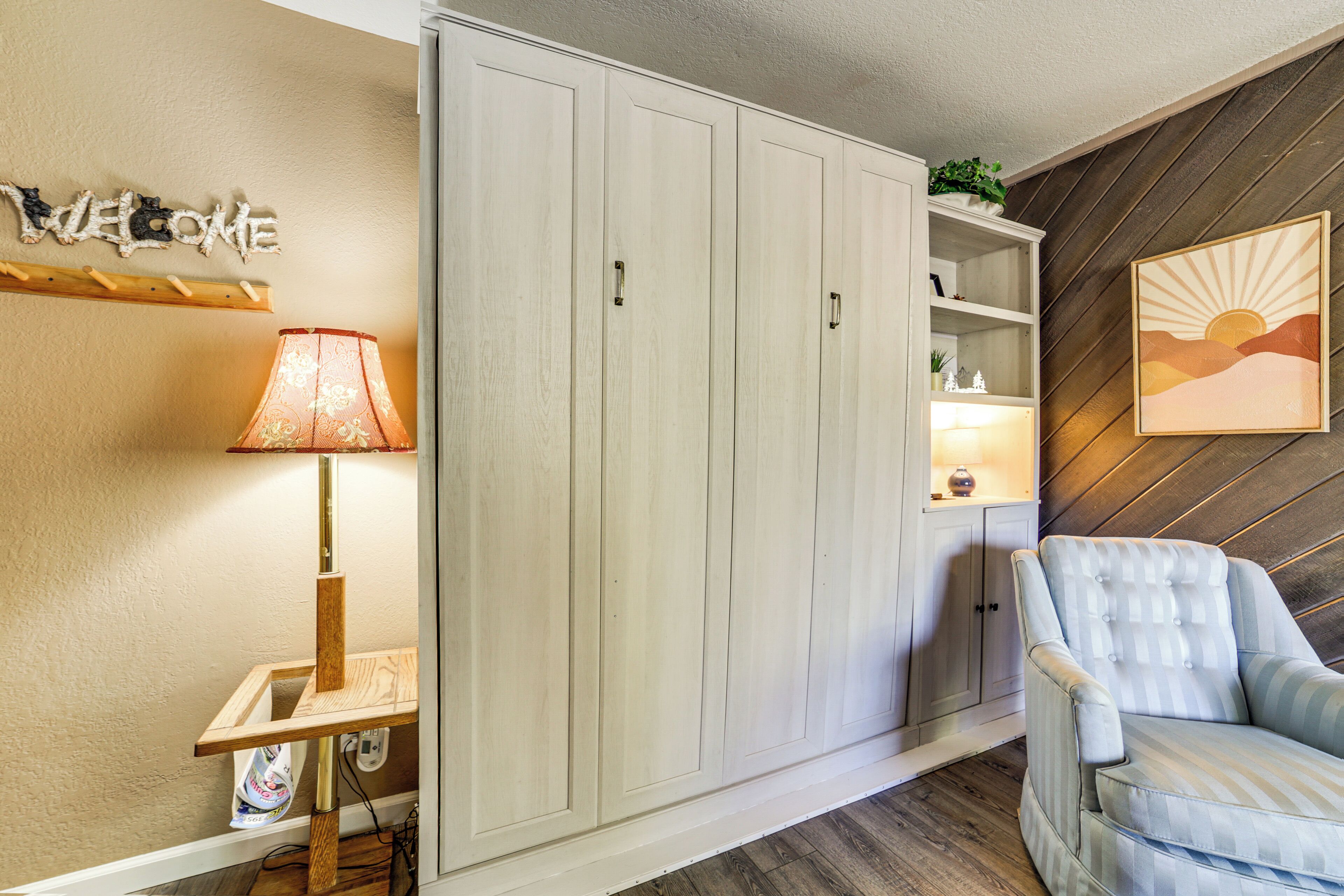 A room with a wardrobe, a lamp, a chair, and a shelf with decorative items.