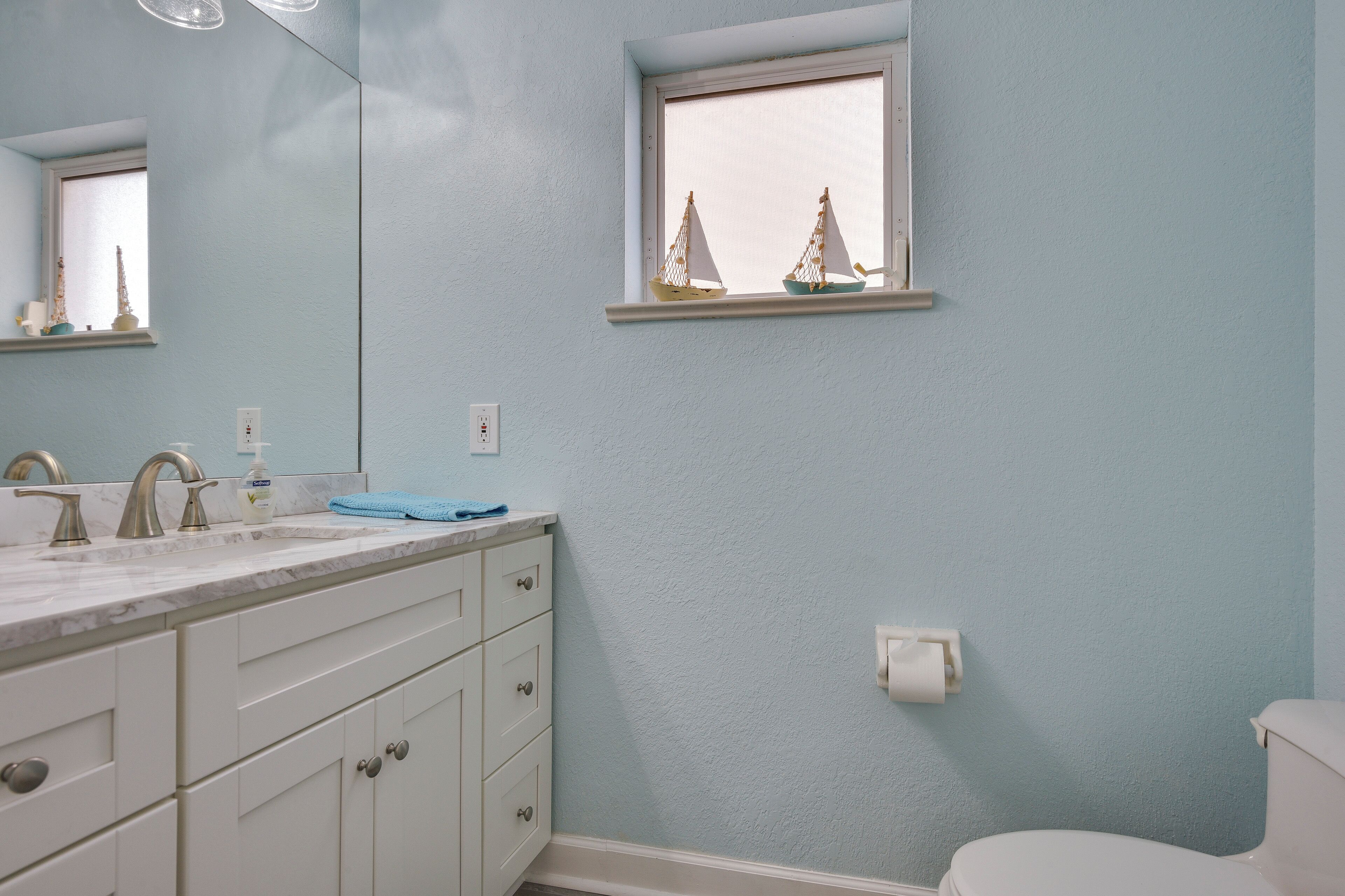 Un bagno con un lavandino bianco, due rubinetti, uno specchio e una finestra decorata con una piccola barca a vela.