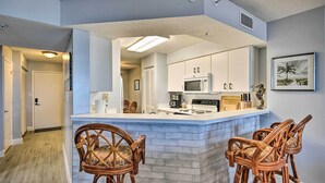 A kitchen with white cabinets, a microwave, and a coffee maker. There are wicker chairs and a white countertop. - Oceanfront Condo Rental Near Daytona Speedway (Ormond Beach)