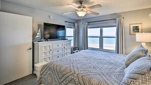 A bedroom with a bed, a ceiling fan, a dresser, a TV, and a window with a view of the beach. - Oceanfront Condo Rental Near Daytona Speedway (Ormond Beach)