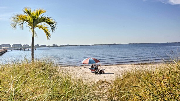 Eine Strandansicht mit einer Palme, einer Person unter einem Sonnenschirm und einer weiter entfernten Stadtansicht.