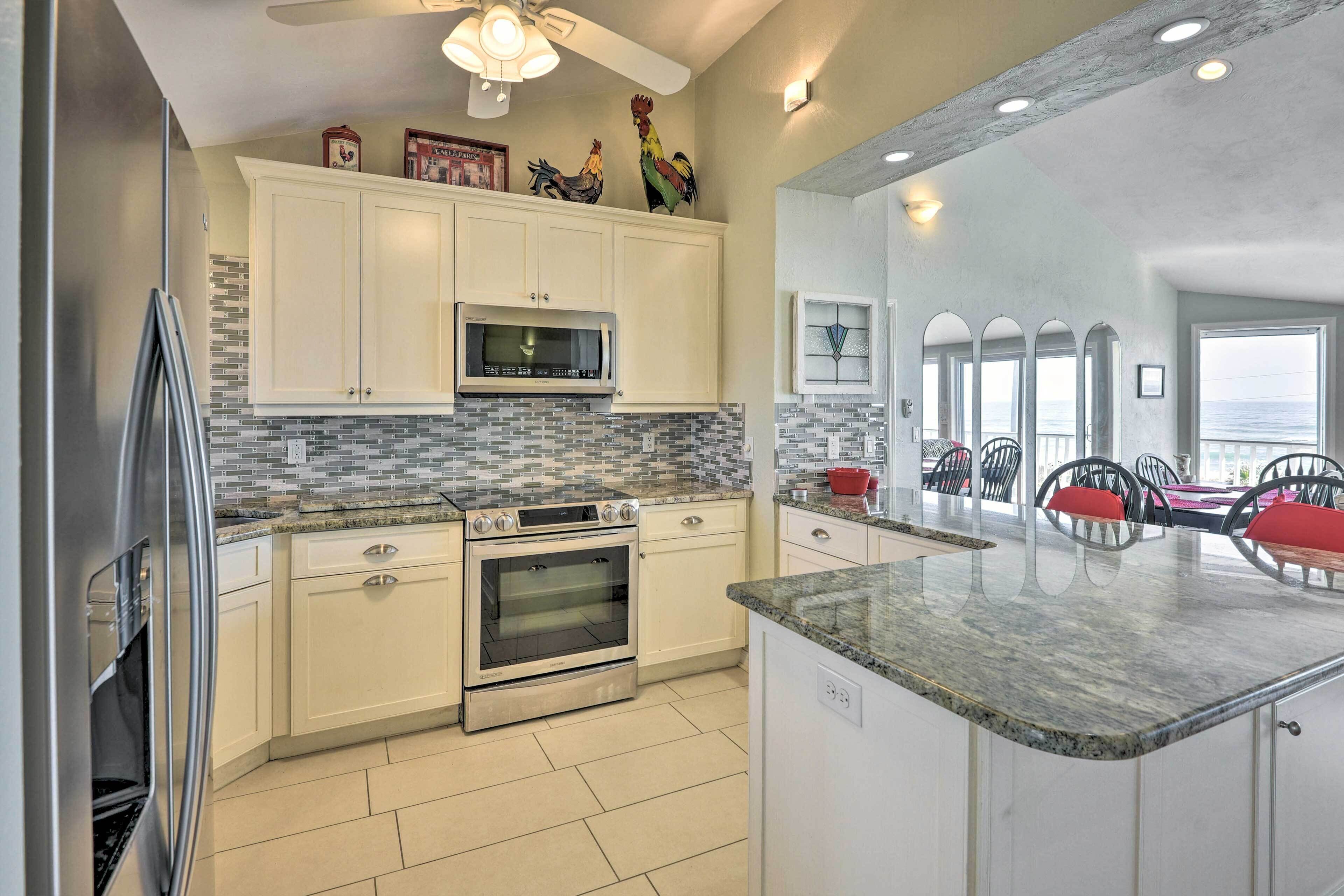A kitchen with white cabinets, a microwave, two ovens, a granite countertop, and a dining area with red chairs.