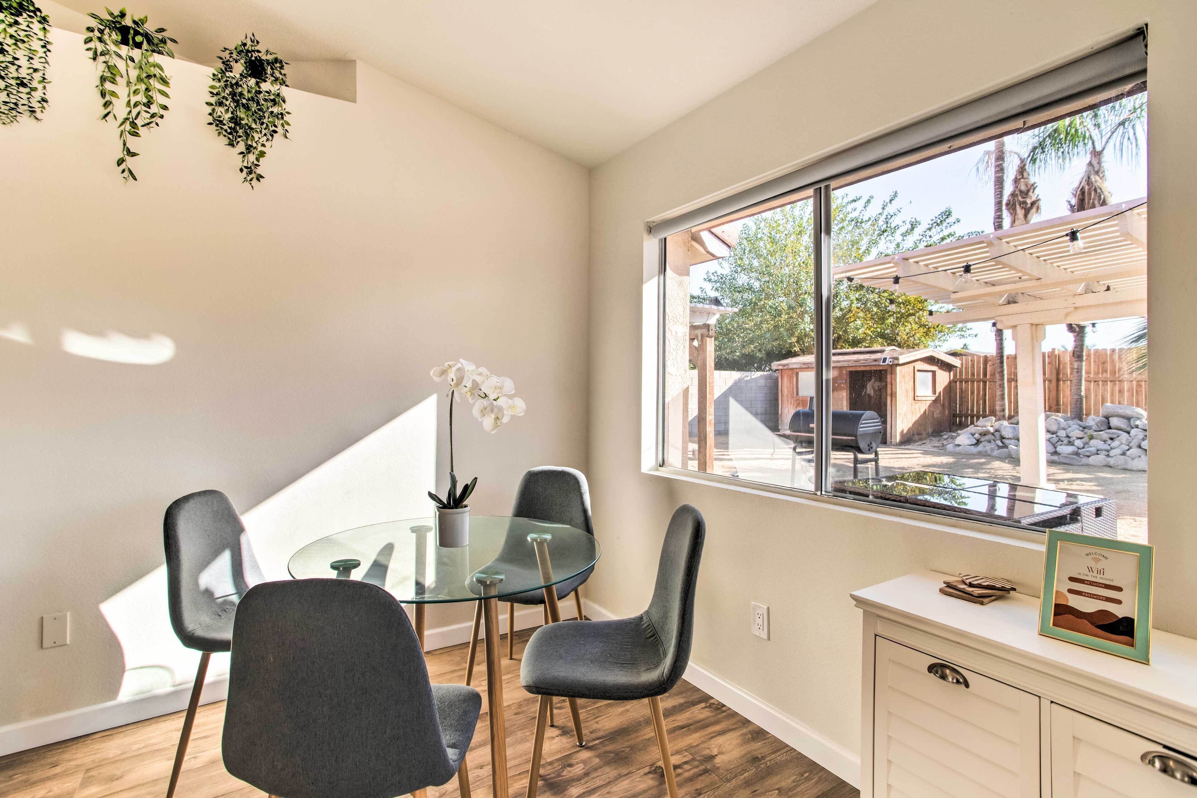 A modern dining area with a glass table, grey chairs, and a view of a backyard through a large window.