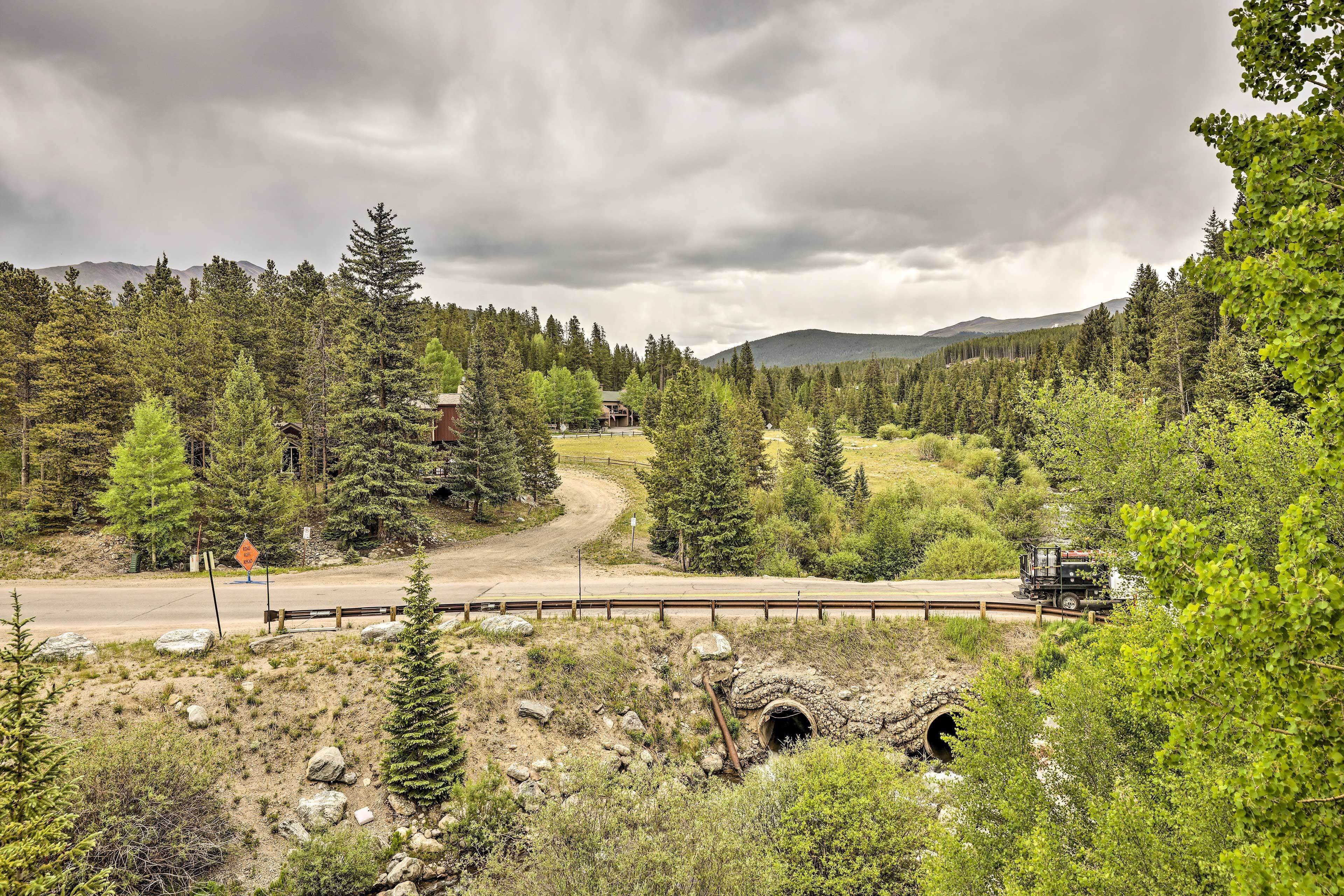 A scenic landscape with a dirt road, a wooden bridge, and a train passing through a forested area with mountains in the background.