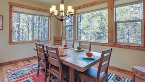 A dining area with a wooden table, chairs, and a chandelier. Large windows with a view of trees are visible. - Beautiful Pagosa Springs Home w/ Deck & Grill (Pagosa Springs)