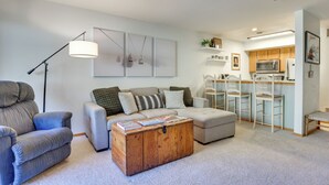 A modern living room with a sofa, armchair, and a wooden coffee table. A kitchen area with a microwave and bar stools is visible in the background. - Walkable Frisco Condo: Mtn Views & Shared Hot Tub! (Frisco)