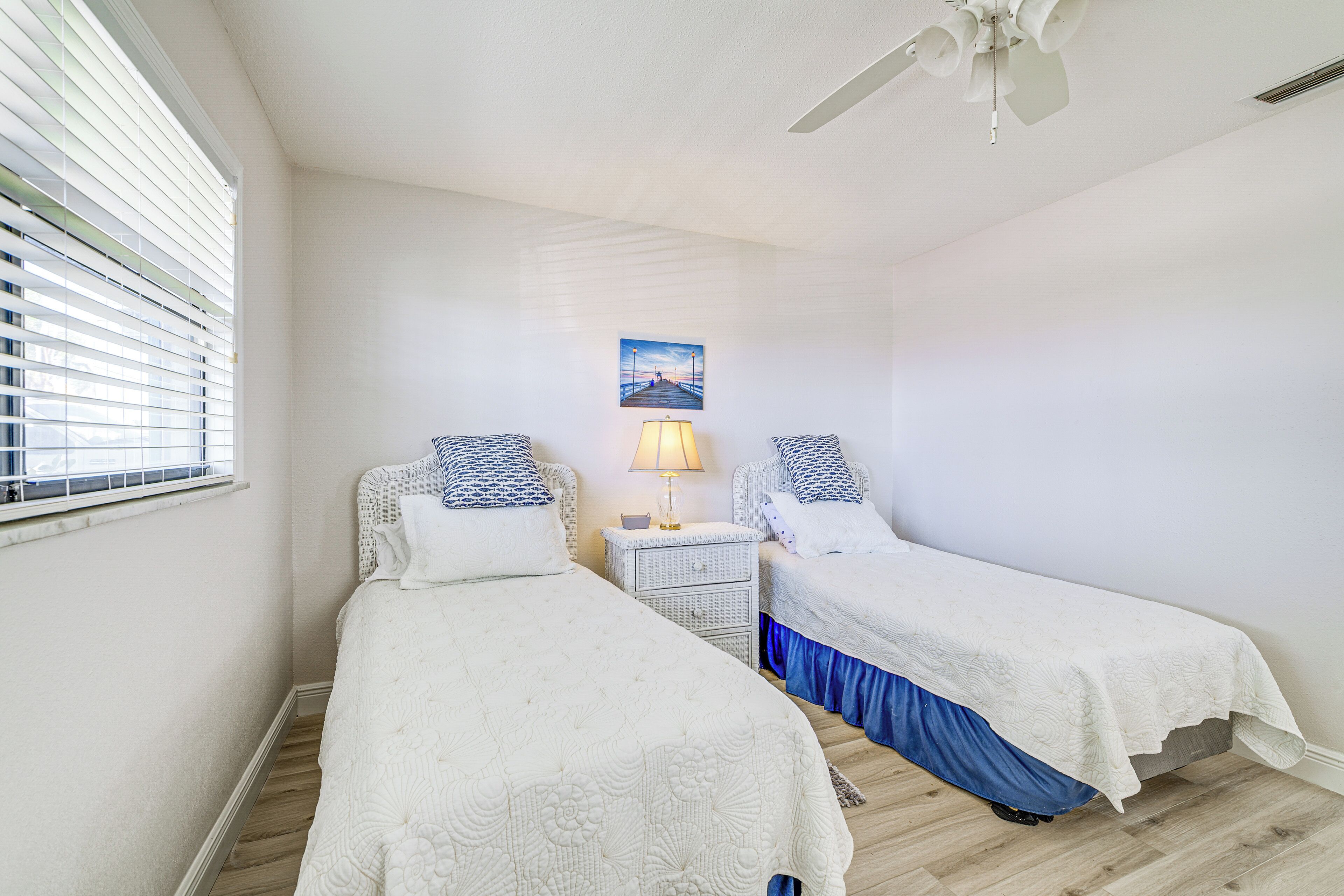 A bedroom with two beds, a nightstand with a lamp, a ceiling fan, and a window with blinds.