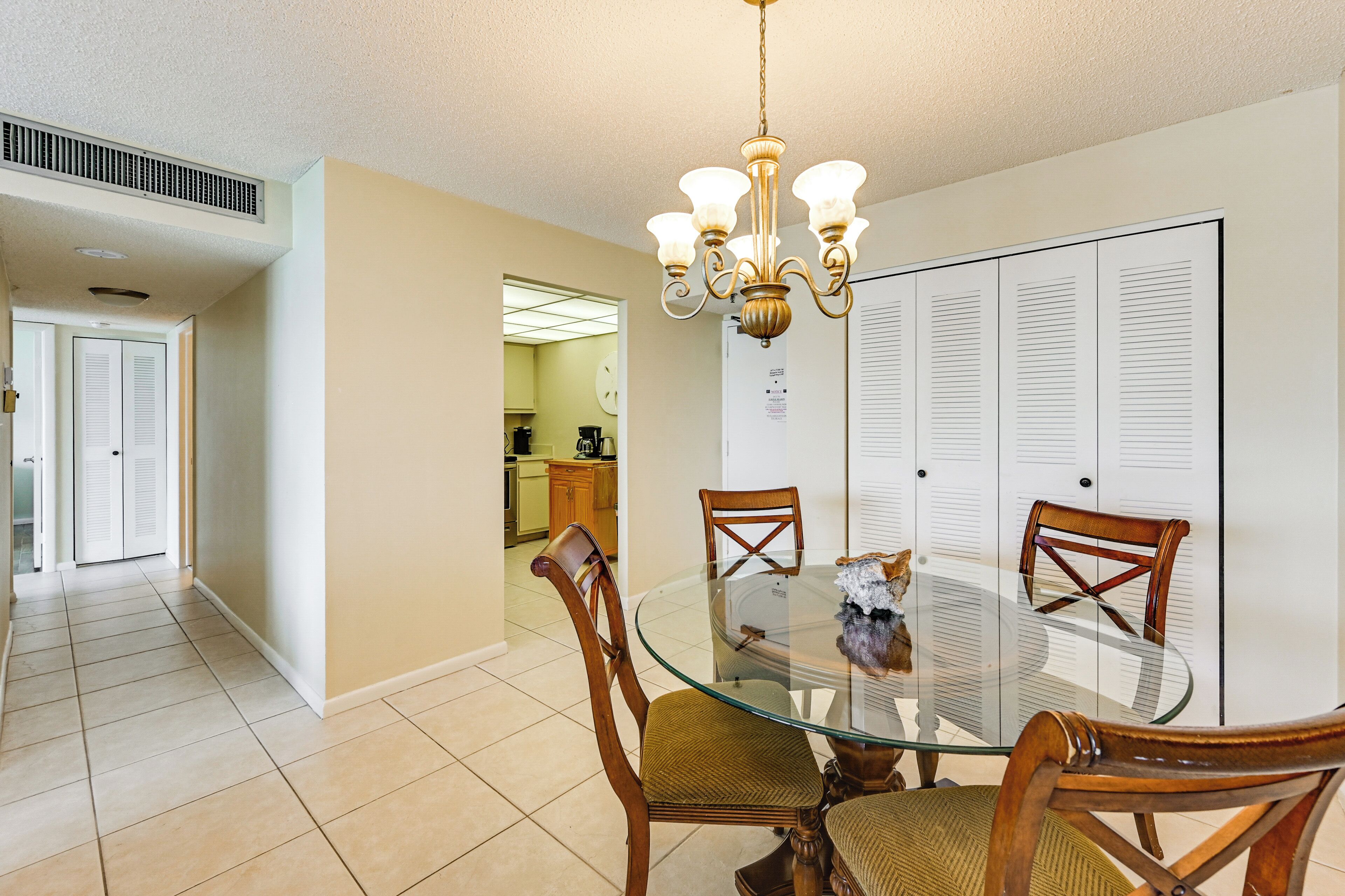 A dining area with a glass table, wooden chairs, a chandelier, and a kitchen in the background.