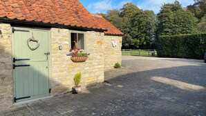 A stone building with a green door, a window with a hanging flower basket, and a small potted plant. - Puddleduck Cottage (Pickering)