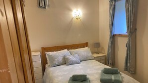 A bedroom with a wooden bed, white linens, a nightstand with a lamp, and a window with curtains. - Puddleduck Cottage (Pickering)