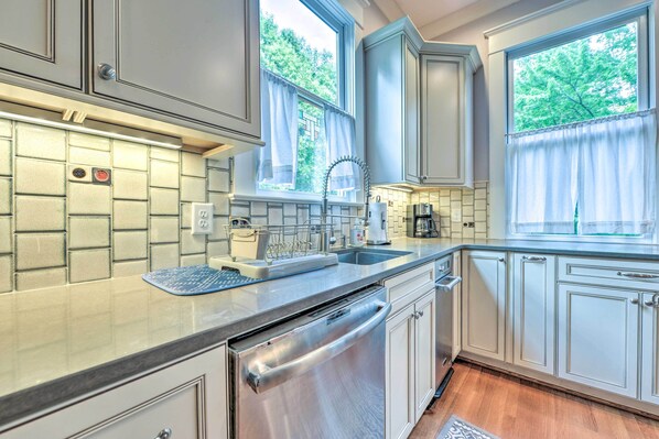 A kitchen with white cabinets, a stainless steel dishwasher, a window with curtains, and a tiled backsplash. - Elegant Raleigh Home w/ Porch, Walk Downtown! (Raleigh)