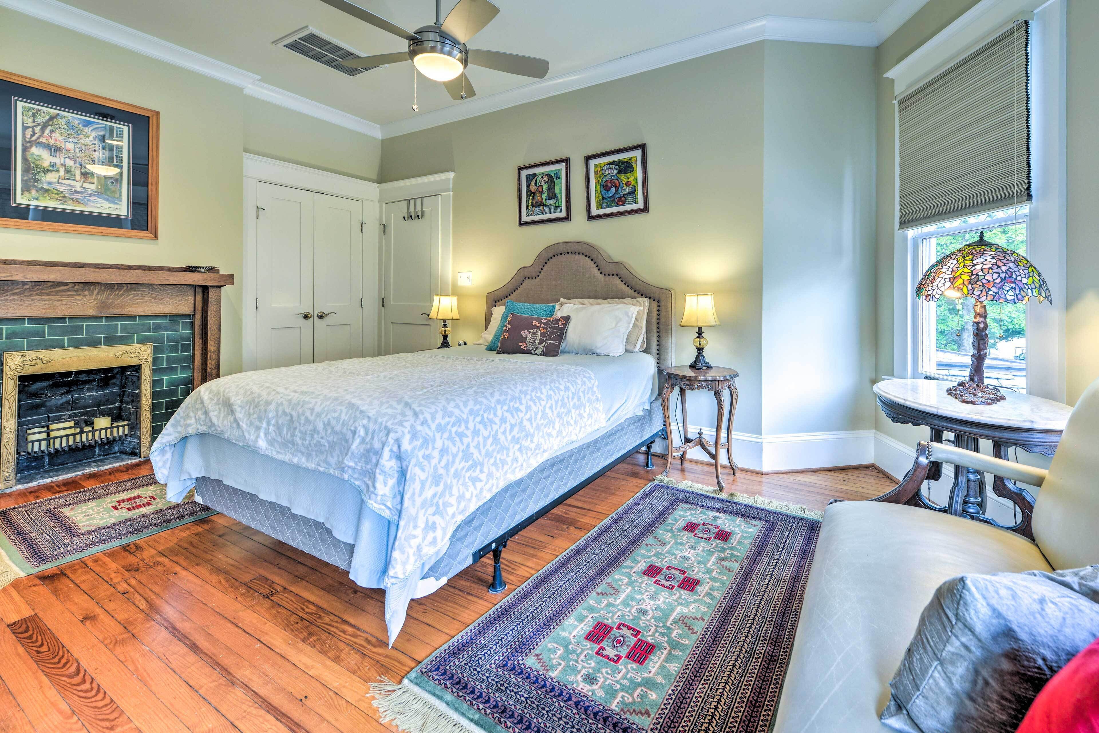 A bedroom with a large bed, two bedside lamps, a fireplace, a window with blinds, and a small table with a lamp.