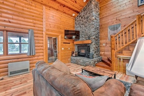 A log cabin interior with a stone fireplace, wooden walls, a sofa, and a staircase.