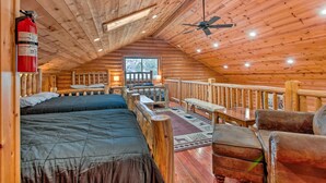 Une chambre située dans une cabane en rondins, comprenant un lit, un bureau, une chaise, un extincteur et un ventilateur de plafond.