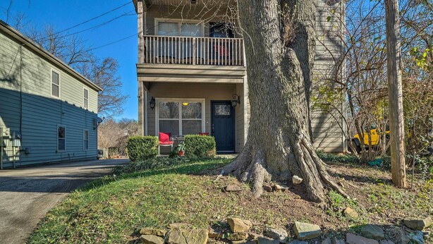 A two-story house with a balcony, a large tree in front, and a yellow vehicle parked nearby.