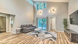 Ein geräumiges Wohnzimmer mit einem grauen Sofa, einem runden Couchtisch und einem geometrischen Teppich. Zwei Fenster mit Vorhängen, eine Tür zum Balkon und eine Pflanze auf der rechten Seite.