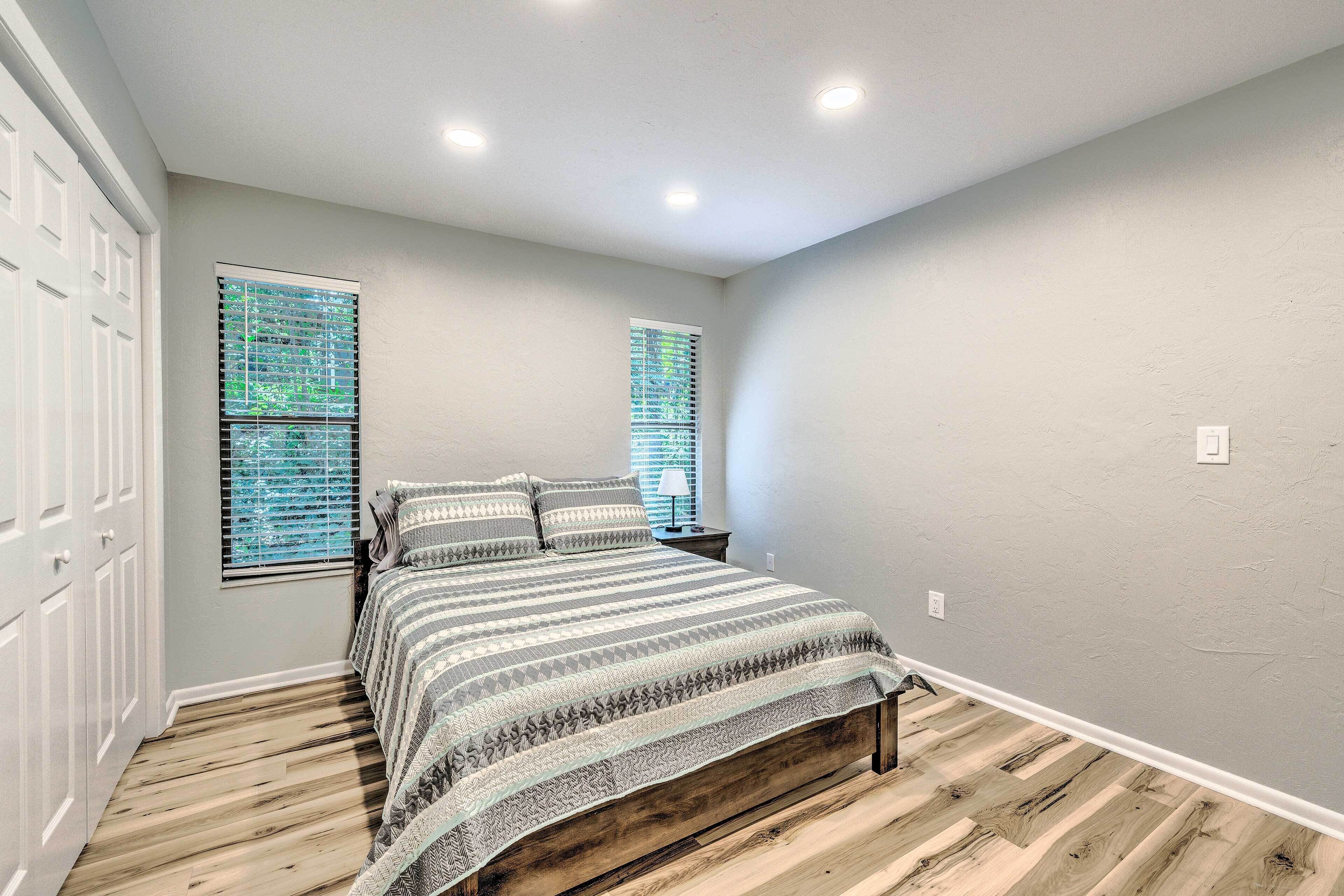 A bedroom with a bed, two windows with blinds, a nightstand, and a wooden floor.