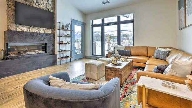 A modern living room with a stone wall, fireplace, leather sofa, and large windows offering a view of mountains. - Condo w/ Mountain Views in Heart of Downtown! (Winter Park)