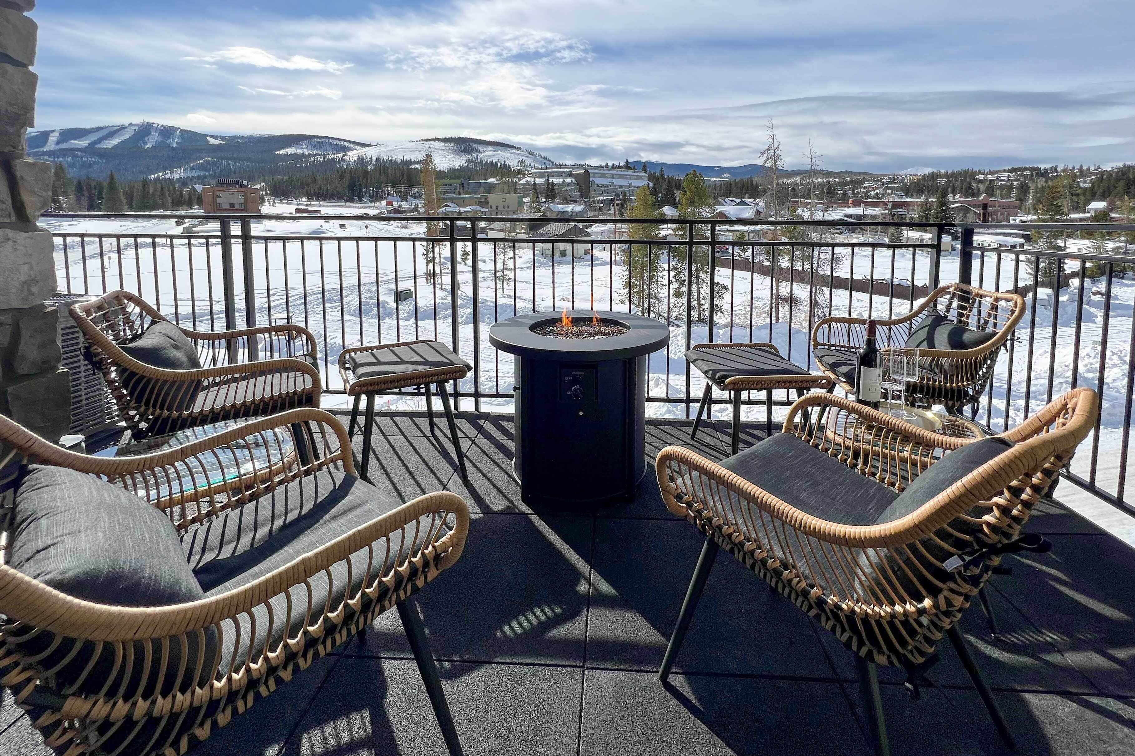A balcony with wicker chairs and a central fire pit, overlooking a snowy landscape with mountains in the distance.