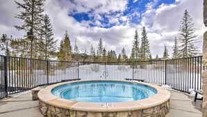 A hot tub surrounded by a stone wall and a black metal fence, with trees and a cloudy sky in the background. - Condo w/ Mountain Views in Heart of Downtown! (Winter Park)