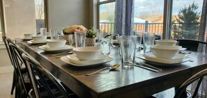 Una mesa de comedor dispuesta con cuencos blancos, platos y vasos transparentes, con vistas a un paisaje montañoso a través de amplias ventanas.